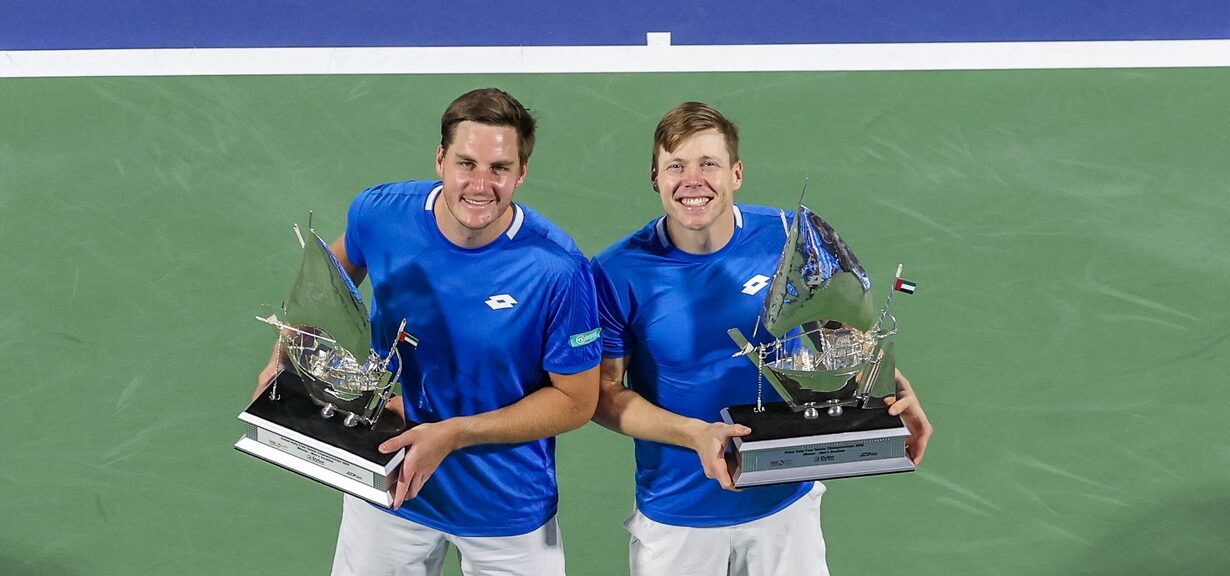 Two tennis players in blue shirts and white shorts stand on a green court, holding silver sail-shaped trophies above a large white "DUBAI" logo.
