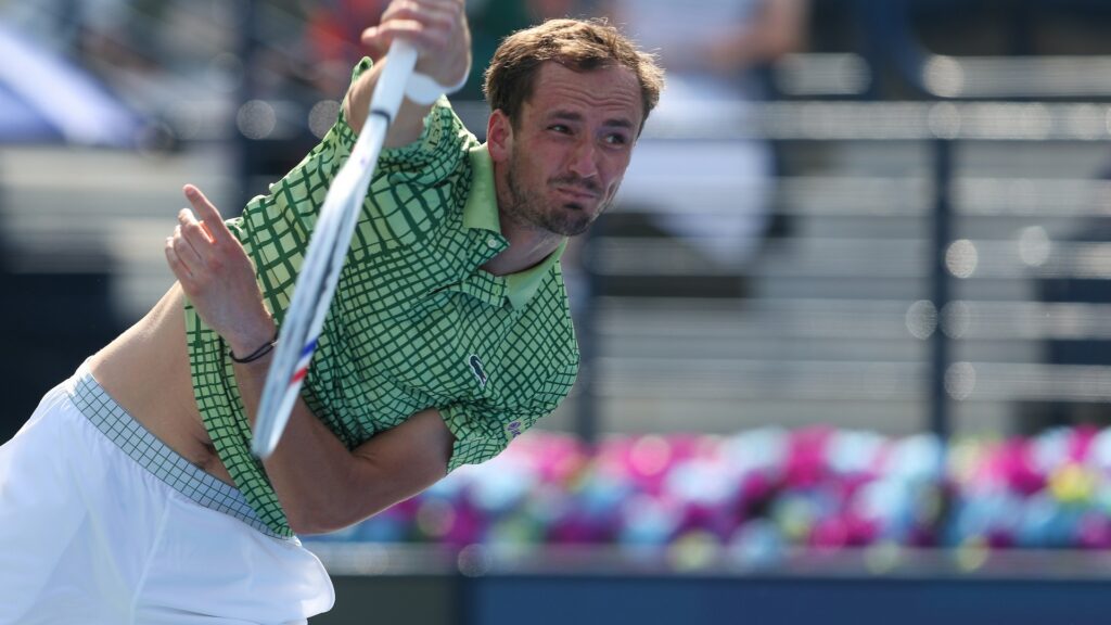 Daniil Medvedev, wearing a green patterned shirt and white shorts, is captured mid-swing on a tennis court, looking intently forward with his shirt slightly lifted.