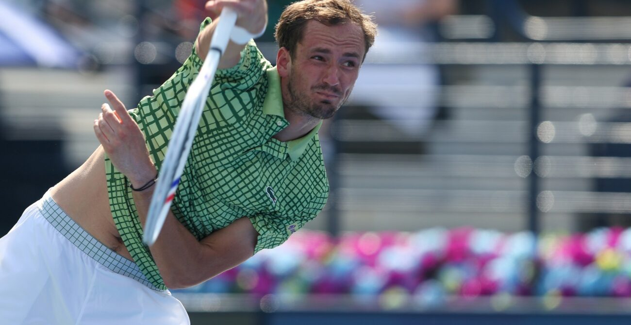 Daniil Medvedev, wearing a green patterned shirt and white shorts, is captured mid-swing on a tennis court, looking intently forward with his shirt slightly lifted.