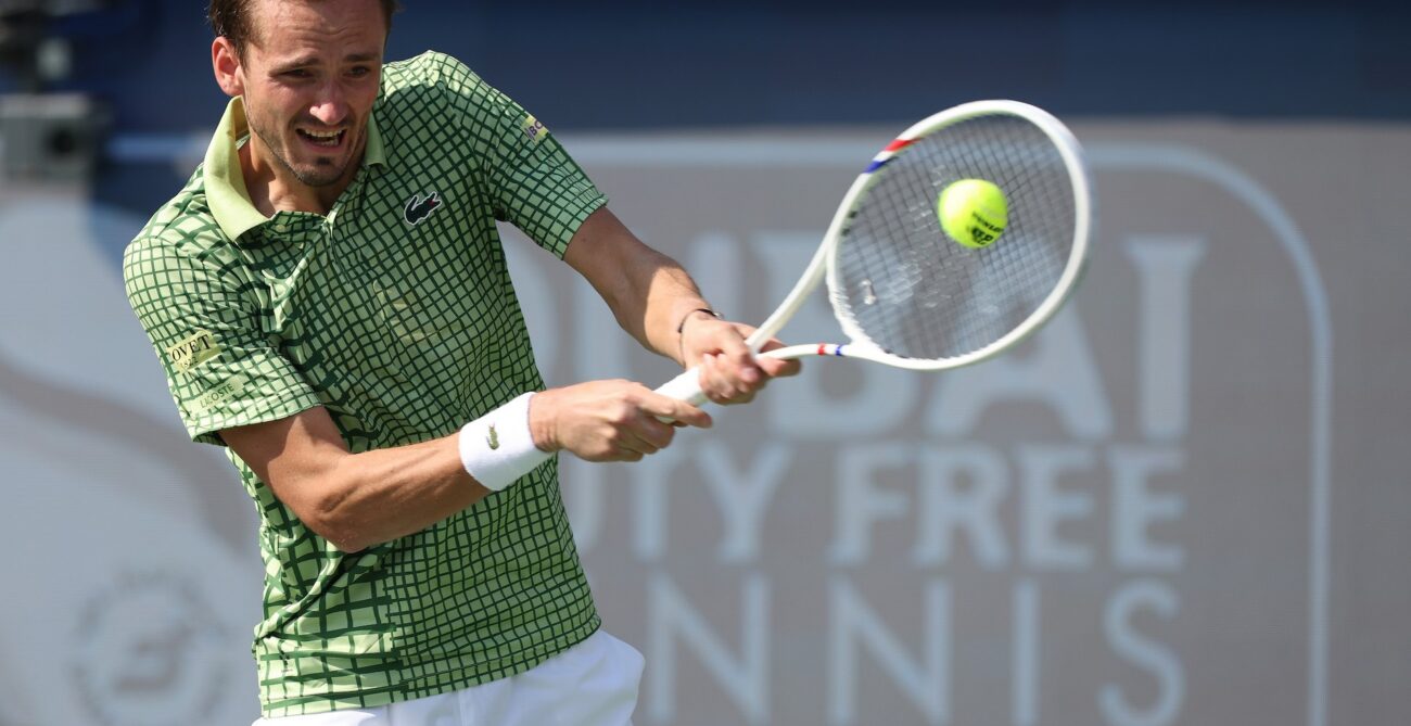 Daniil Medvedev, in a green patterned shirt and white shorts, intensely hits a tennis ball with his racket during a match.