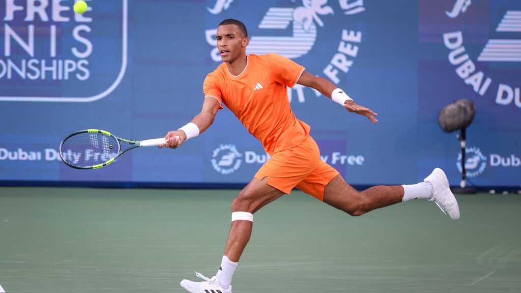 Felix Auger-Aliassime lunges across a green court to hit a forehand shot, dressed in a bright orange tennis kit. The match is set against a blue background featuring "Dubai Duty Free" logos.