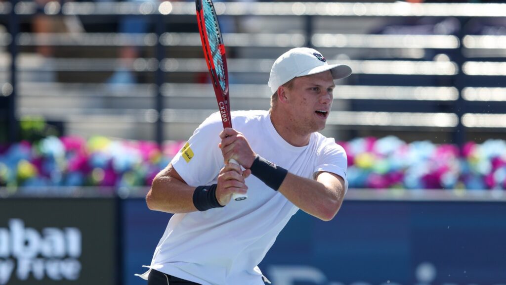 Mid-action, Jenson Brooksby plays tennis, focused and holding a red and black racket. He wears a white cap and t-shirt on a sunny outdoor court.