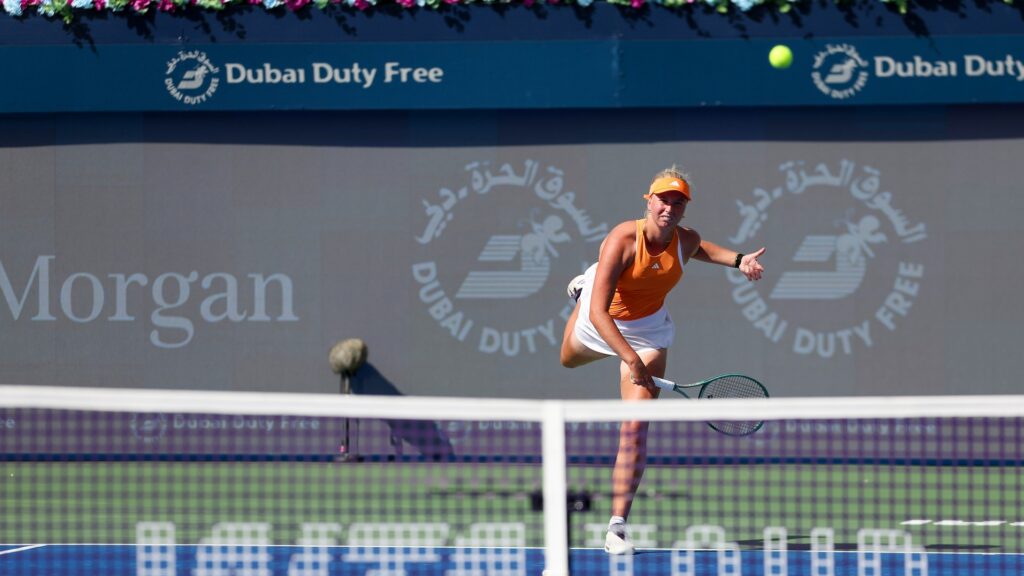 Clara Tauson lunges forward to strike a tennis ball during a match on a blue court. She wears an orange top and white skirt, with "Dubai Duty Free" branding and flower arrangements in the background.
