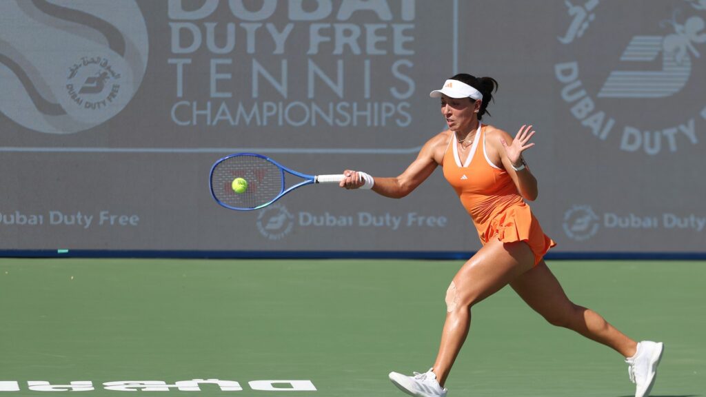 Jessica Pegula, in an orange outfit, lunges forward to hit a tennis ball with her blue racket on a green court. A "Dubai Duty Free Tennis Championships" banner is behind her.