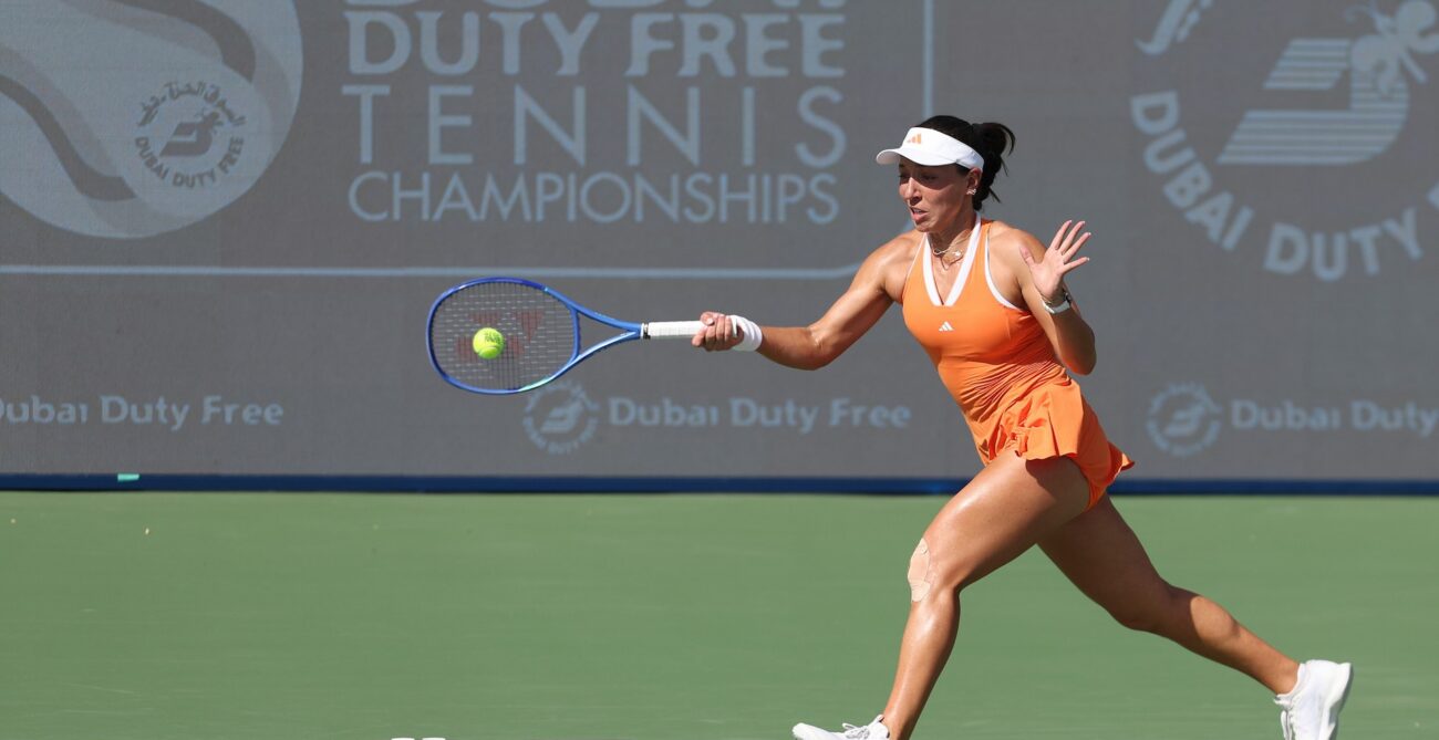 Jessica Pegula, in an orange outfit, lunges forward to hit a tennis ball with her blue racket on a green court. A "Dubai Duty Free Tennis Championships" banner is behind her.