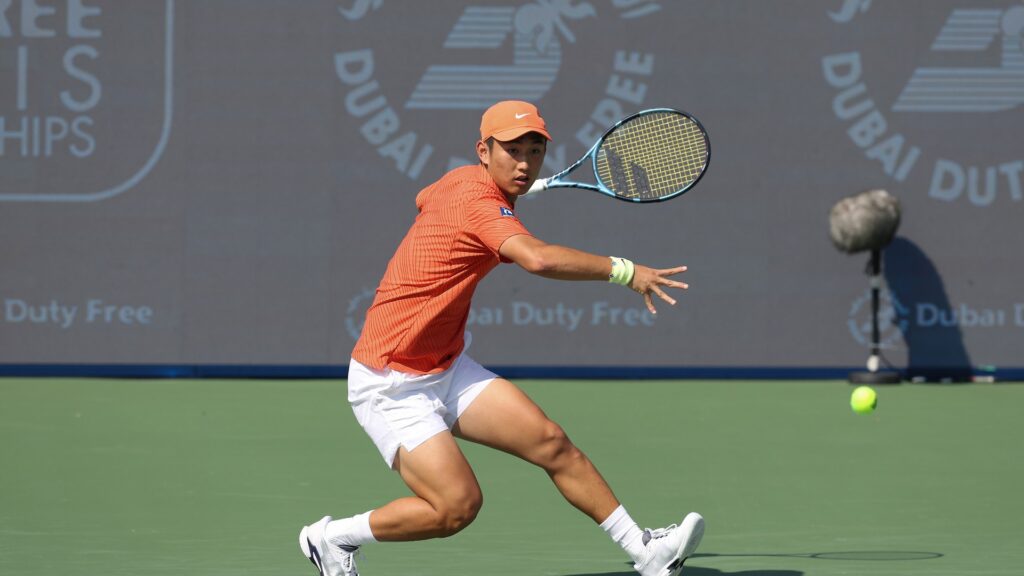 Shang Juncheng lunges forward on a tennis court, preparing to hit a tennis ball with his racket. He wears an orange shirt, white shorts, and an orange cap, with "Dubai Duty Free" banners visible in the background.