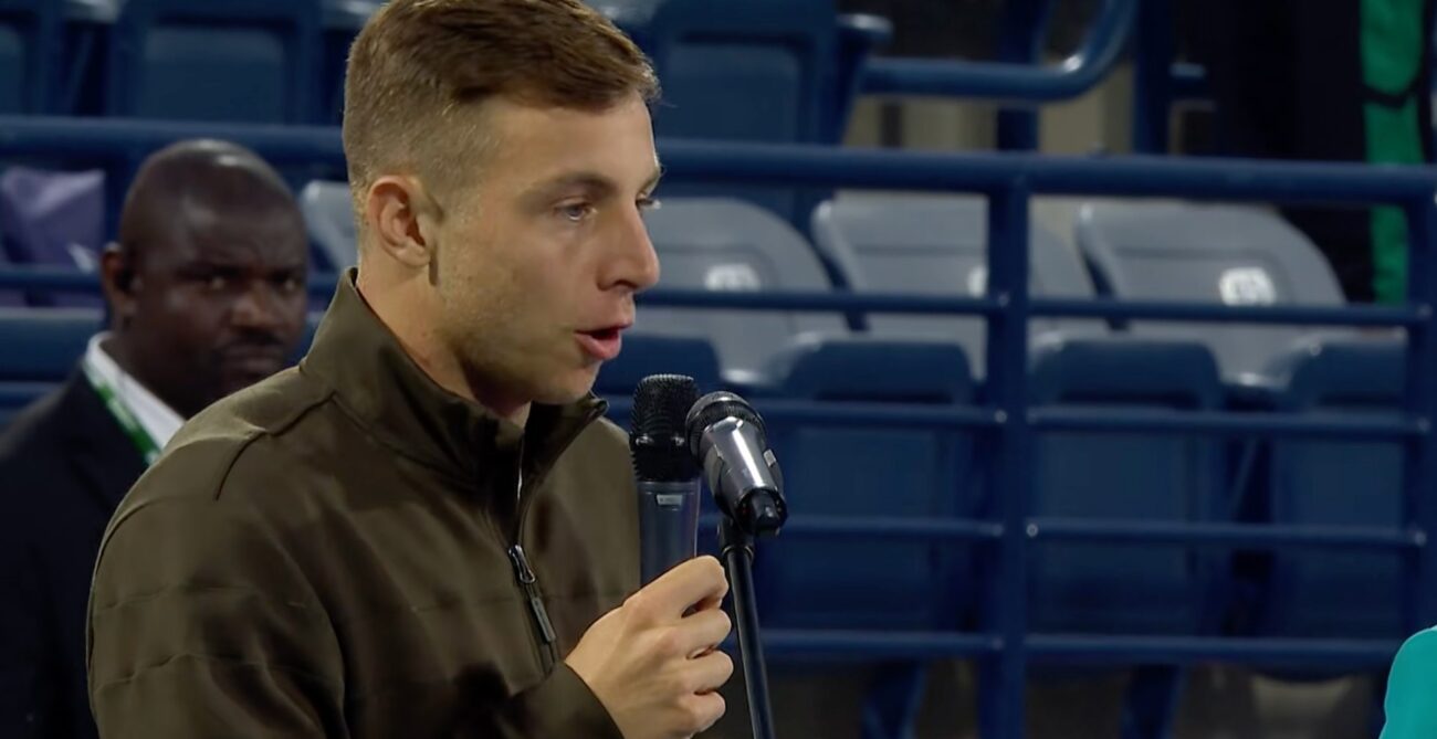 Tallon Griekspoor speaks into a handheld microphone while wearing an olive green jacket, with blue stadium seating in the background.