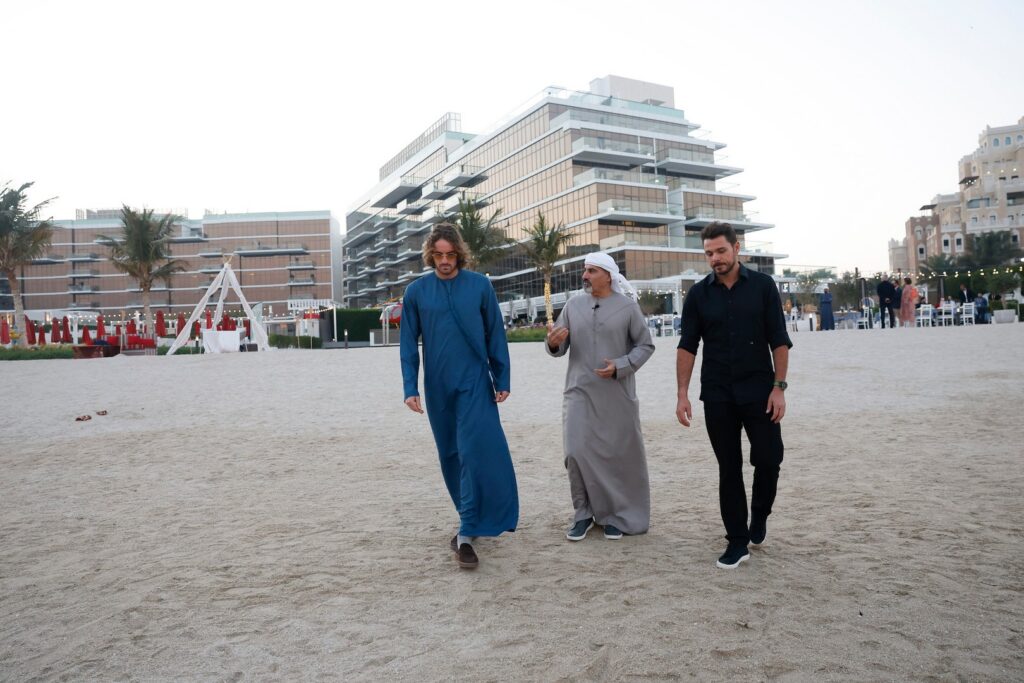 Stan Wawrinka and Stefanos Tsitsipas walk with another Stan Wawrinka and Stefanos Tsitsipas on a sandy area in front of modern buildings. Palm trees and outdoor furniture are visible in the background.