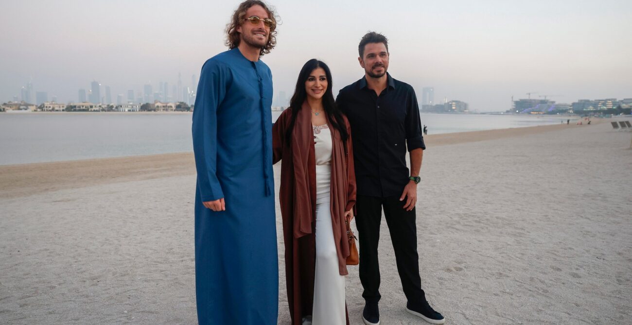 Stan Wawrinka and Stefanos Tsitsipas stand on a beach, with one wearing a blue garment. A third Stan Wawrinka and Stefanos Tsitsipas is with them, and the cityscape is visible in the background.