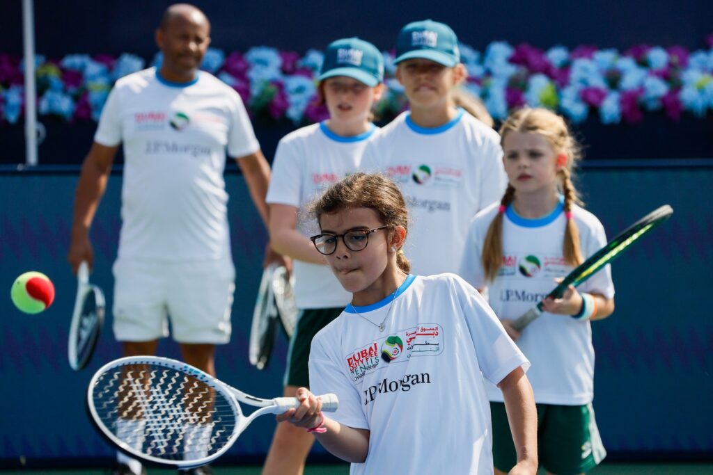 Participants playing tennis, with a focus on a young player in glasses preparing to hit a ball. Others in the background wear matching sports attire and watch the play.