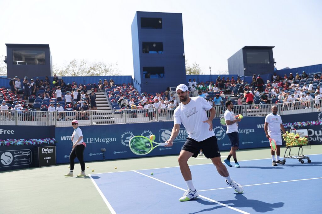 Participants play tennis on a court surrounded by spectators in the stands. One participant is actively hitting a ball with a racquet.