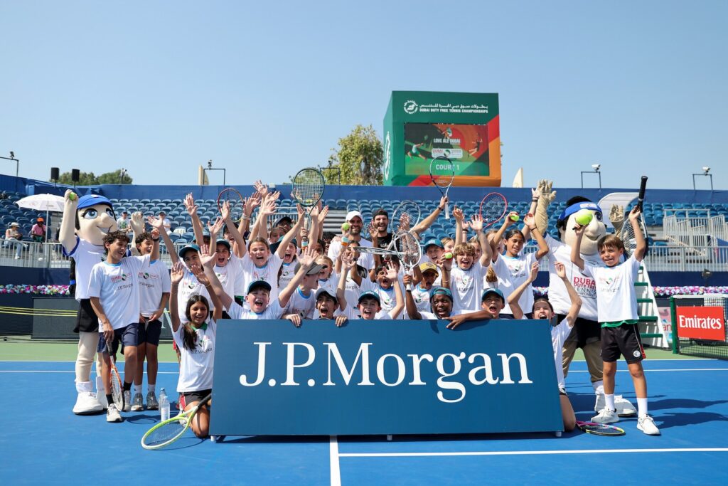 Participants joyfully gather on a tennis court, holding rackets and raising their arms, with large JP Morgan signage in front and a tennis mascot on the side.